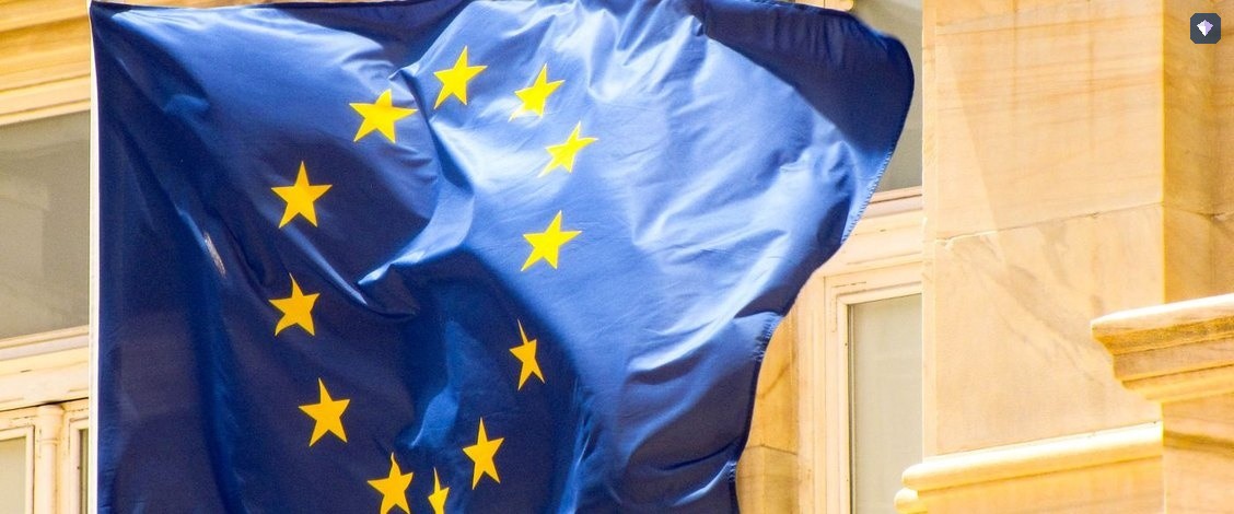 The blue and gold flag of the European Union flying beside a stone building