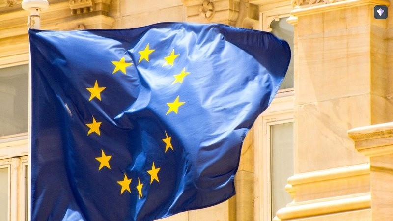 The blue and gold flag of the European Union flying beside a stone building