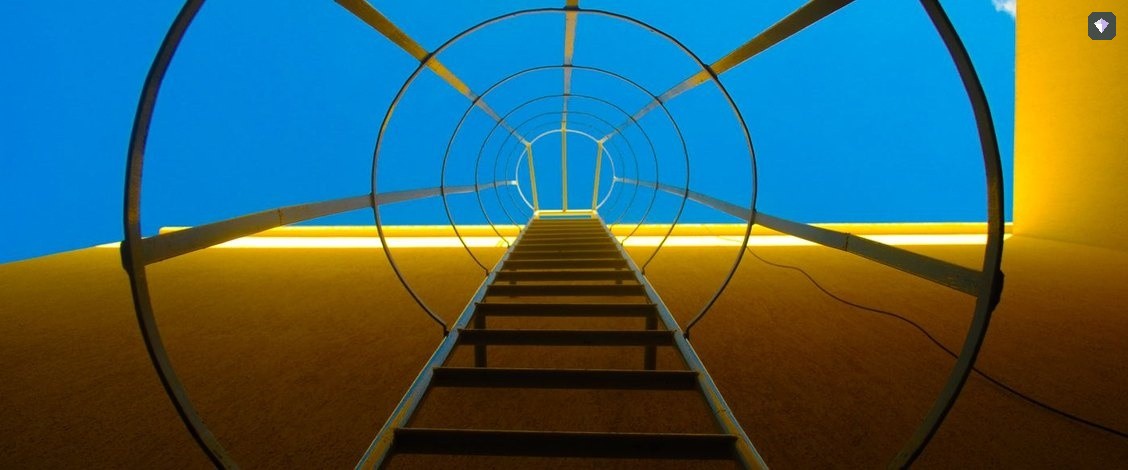 A steel ladder curving up a yellow wall against a blue sky