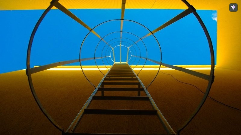 A steel ladder curving up a yellow wall against a blue sky