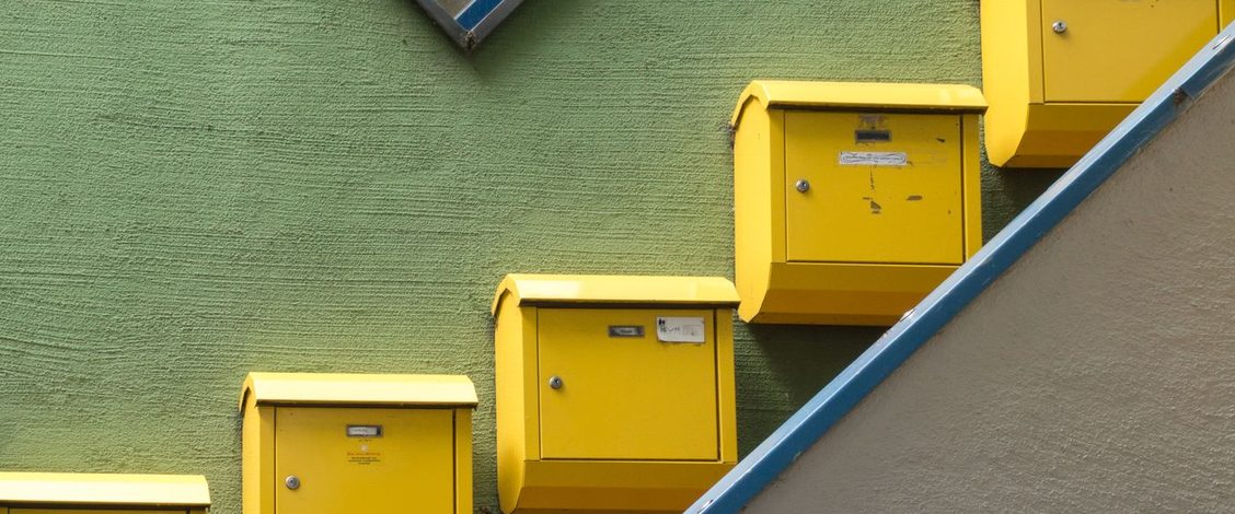 Yellow letterboxes climbing a green rendered wall beside a stair rail painted blue