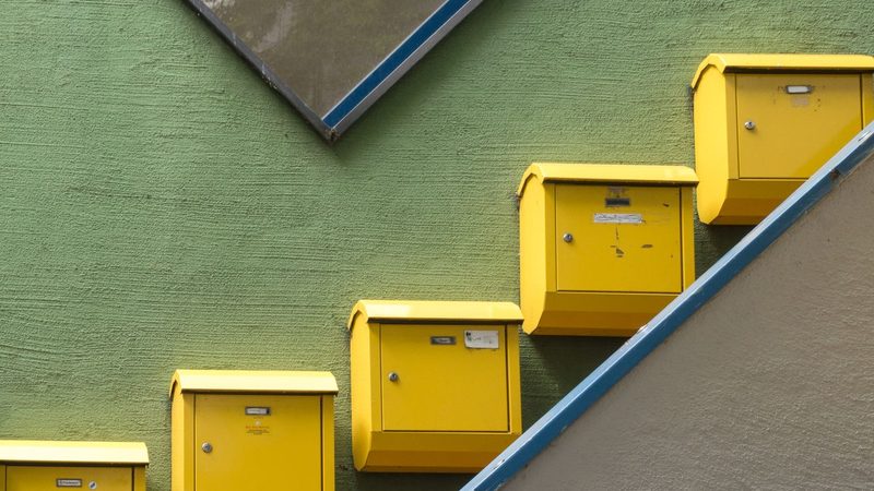 Yellow letterboxes climbing a green rendered wall beside a stair rail painted blue