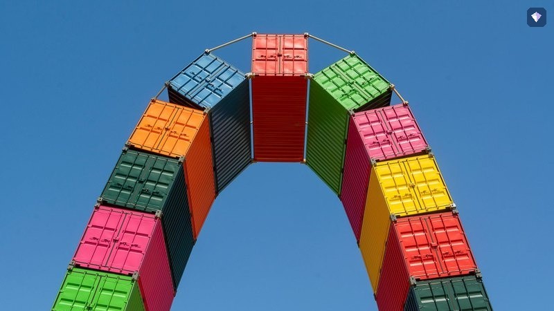 Shipping containers in many colours stacked into an arch against a blue sky
