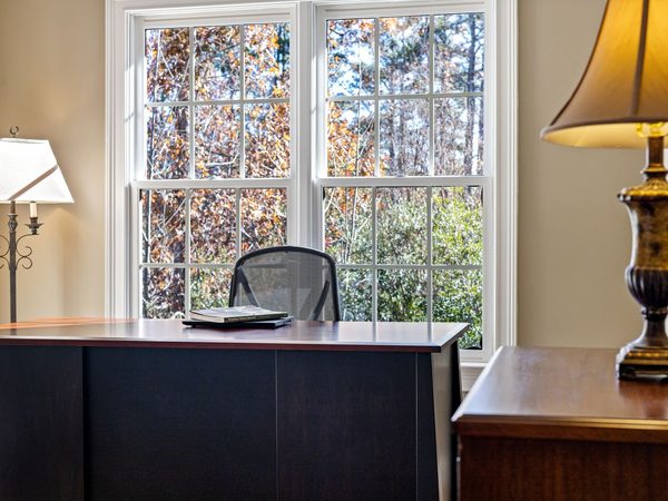 A desk set by a tall sash window with a lamp on the sideboard and an empty chair