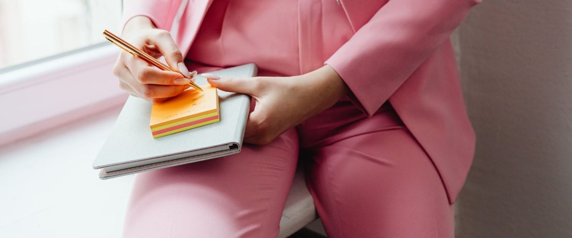 A woman in a pink trouser suit sits on a bright windowsill with a grey notebook on her knee, writing on an orange and yellow pad of sticky notes with a gold pen