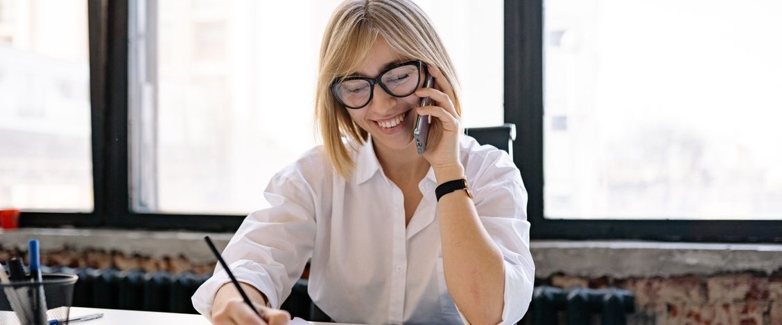 A fee earner in a white shirt and glasses laughing on a phone call while she writes a note on a clipboard, at a white desk by tall windows with an exposed brick wall behind her